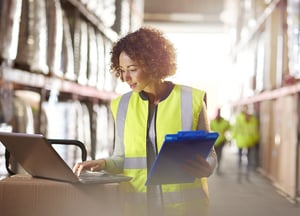 Woman in a yellow safety vest looking at a laptop inside a warehouse